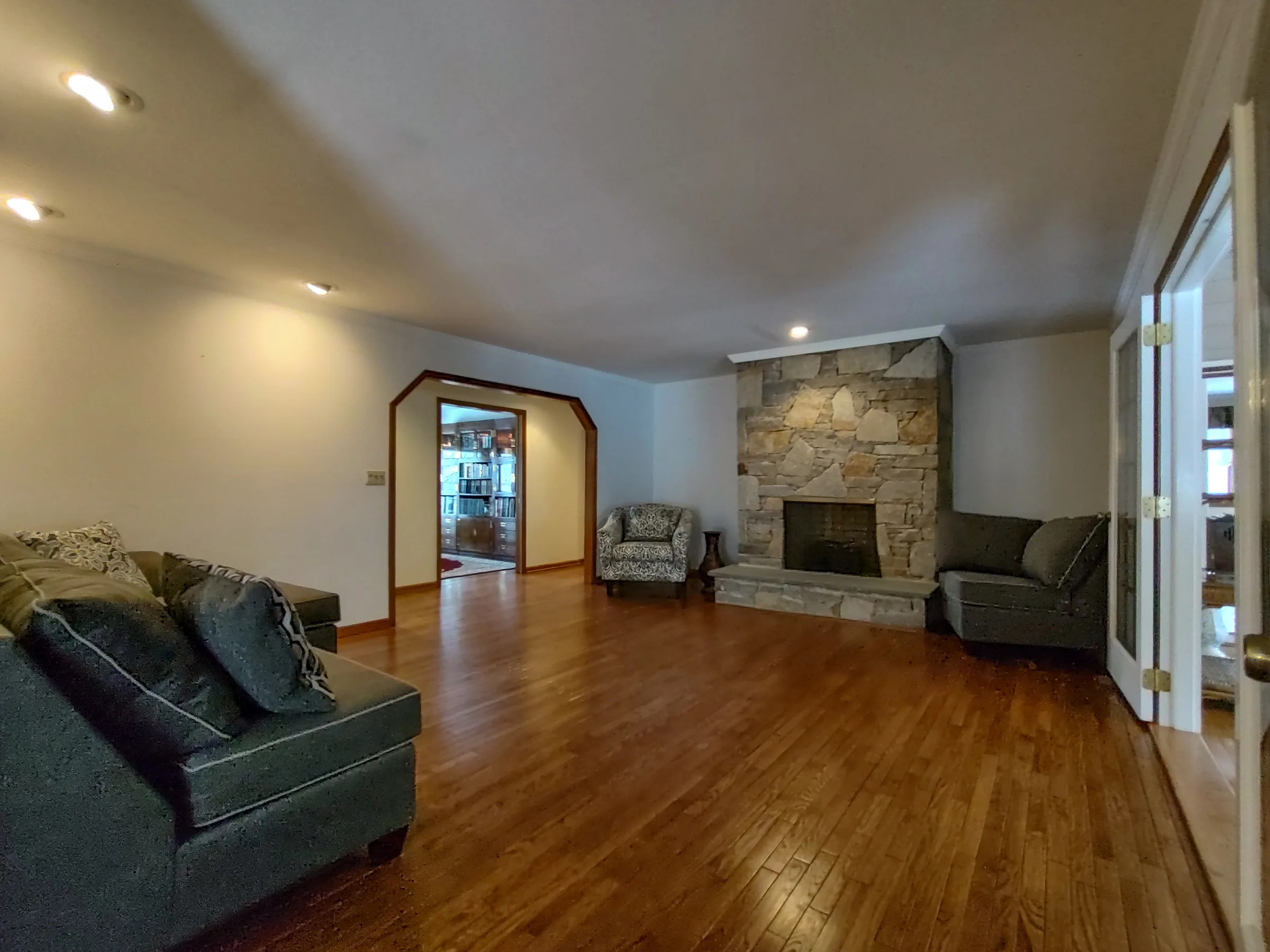 Spacious living room with a full floor-to-ceiling stone fireplace, warm hardwood floors, and an arched doorway opening to a book-lined study
