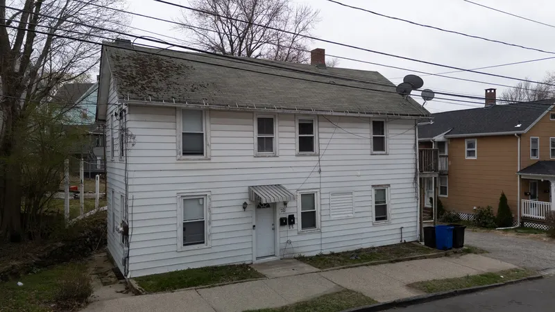 Low aerial of a two-story white clapboard home with a striped door awning, set among neighboring houses